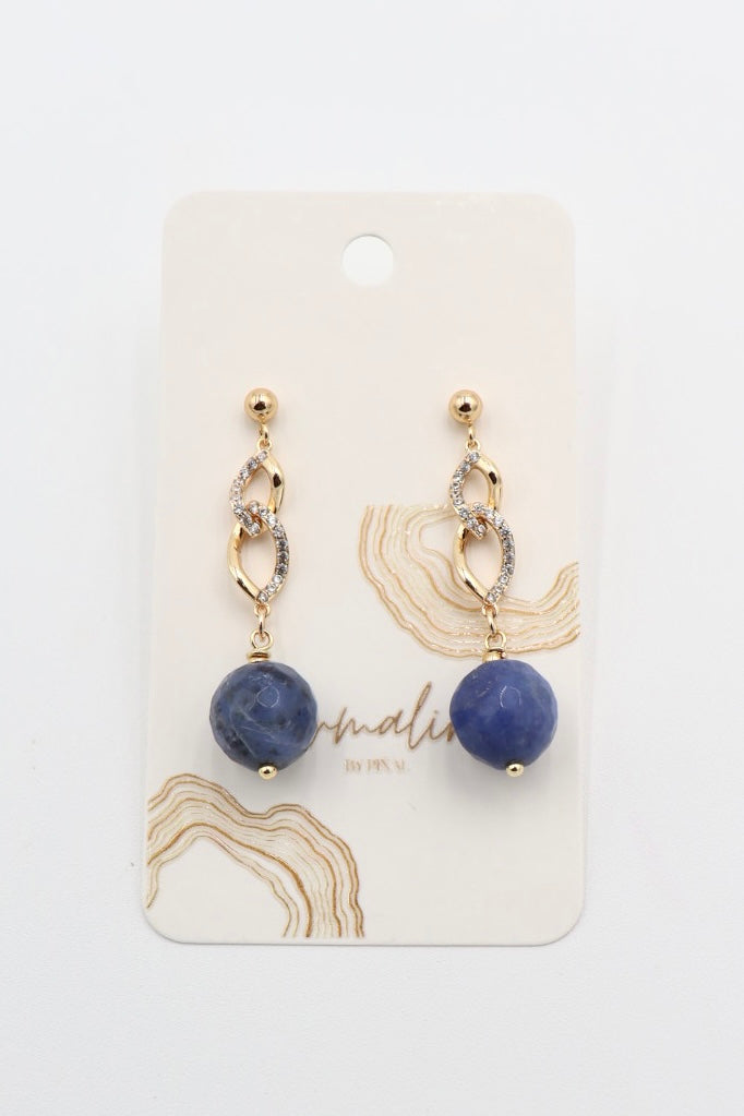 A pair of gold drop earrings with a textured gold chain, a small clear crystal accent, and a round blue stone sodalite faceted bead dangle, displayed on a cream-colored jewelry card with gold abstract designs.