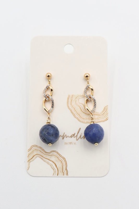 A pair of gold drop earrings with a textured gold chain, a small clear crystal accent, and a round blue stone sodalite faceted bead dangle, displayed on a cream-colored jewelry card with gold abstract designs.